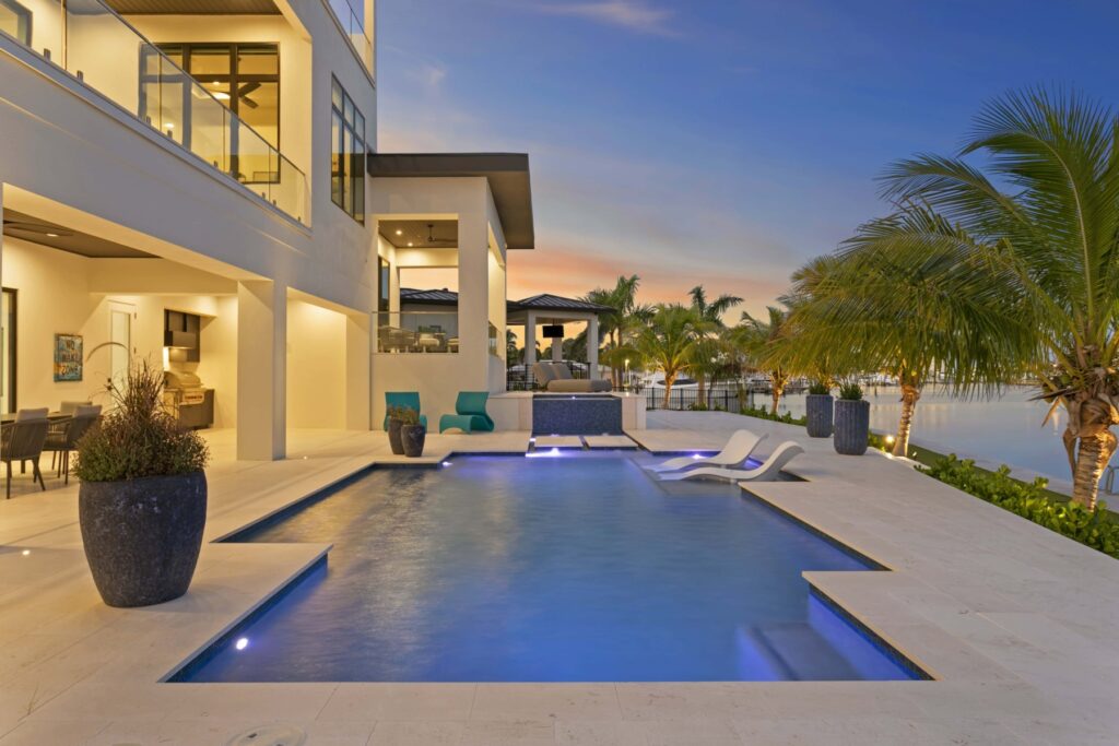 Illuminated pool and loungers beside a modern waterfront home at dusk