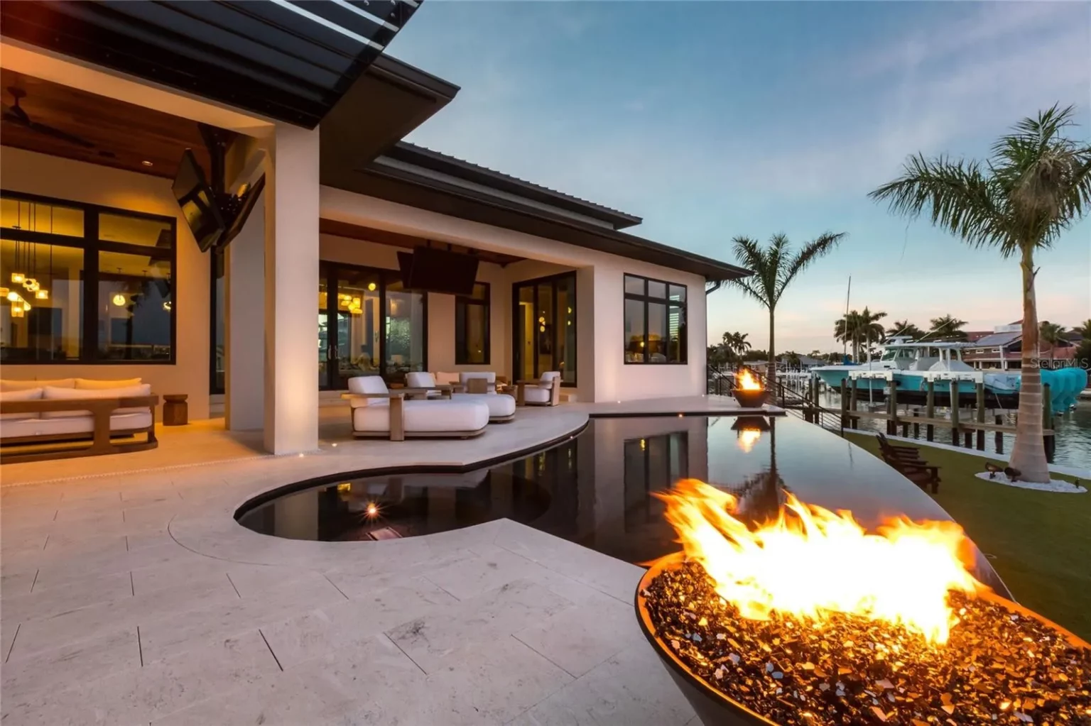 Covered lanai with a linear fire feature overlooking a pool and boat dock at dusk